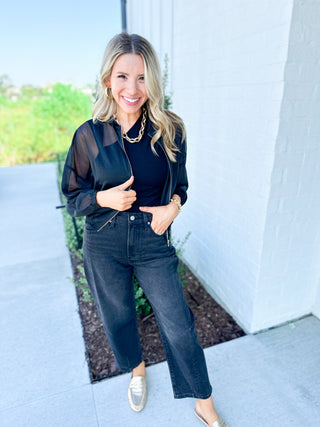 Woman wearing a black outfit with sheer sleeves and gold necklace, standing outdoors.