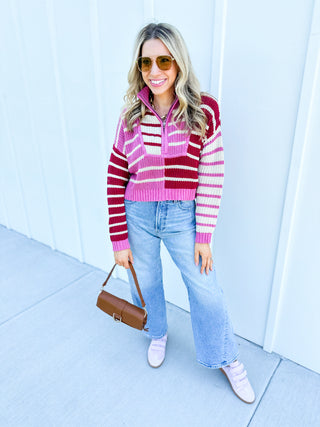 Woman wearing a pink and red striped sweater with blue jeans, standing against a light blue wall.