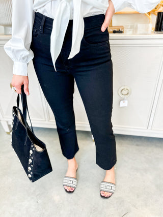 Person wearing black pants and a white top, holding a black handbag indoors.