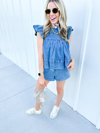 Girl standing in front of a white wall, wearing a denim top and denim short set.