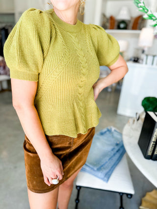Person wearing a green cable knit sweater and brown skirt in an indoor setting.