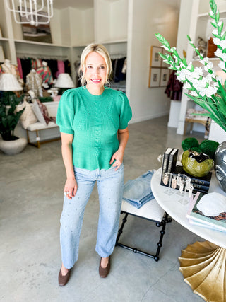 Woman in a green sweater and light blue jeans standing in a stylish room with decor elements.