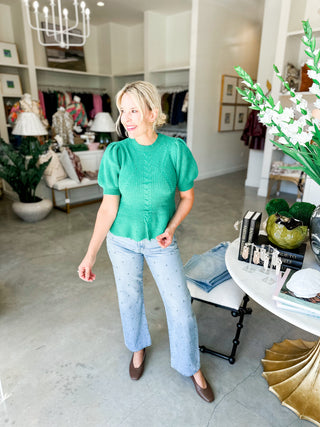 Woman in a green top and light blue jeans standing in a store with decor and furniture.