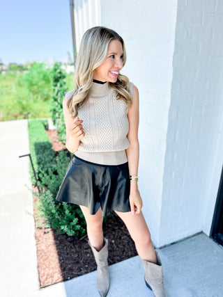 Woman wearing a beige sleeveless top and black leather skirt standing on a porch.