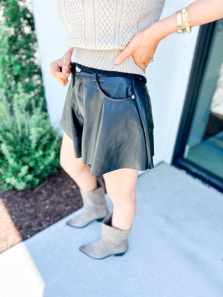 Person wearing a white textured top, dark black leather shorts, and gray ankle boots on a light blue surface.