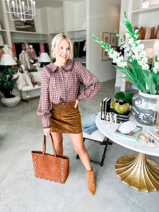 Woman in a plaid shirt and brown skirt holding a brown tote bag in a stylish room.