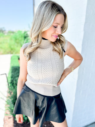 Woman wearing a beige sleeveless sweater and black leather shorts outdoors.