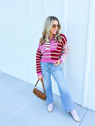 Woman wearing a striped sweater and jeans, holding a brown bag against a light blue wall.